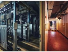 Blick in einen technischen Maschinen- und Schaltraum mit Metallrahmen, Kabelbündeln und großen Elektro-Schaltschränken auf der linken Seite. Rechts verläuft ein schmaler Flur mit rotem Boden, Deckenleitungen und einer Türöffnung mit gelb-schwarzer Warnmarkierung.