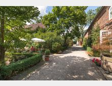 Gepflasterter Weg in einem Außenbereich mit Bäumen und Sträuchern, flankiert von roten Backsteingebäuden. Links befinden sich Tische mit Sonnenschirmen in einem Gartenbereich.