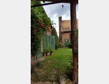 Ein kleiner Hinterhof mit Rasen, umgeben von Backsteingebäuden mit grünen Holztüren. An den Wänden wachsen Kletterpflanzen, und vor den Türen stehen Blumentöpfe; im Hintergrund ist ein Gebäude mit einem roten Ziegeldach zu sehen.