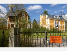Zwei historische Villen mit verzierten Fassaden, eine in rotem Ziegel und die andere in gelbem Putz mit weißen Akzenten, umgeben von Bäumen und Rasenflächen. Im Vordergrund steht ein schmiedeeisernes Tor mit einem Warnhinweis.