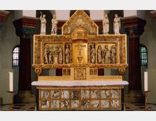 Ein reich verzierter, goldener Altar mit geschnitzten Figuren und religiösen Szenen, umgeben von kleinen Fenstern und hohen Säulen im Hintergrund. Der Altaraufbau zeigt detaillierte Reliefs, und davor steht ein mit weißem Tuch bedeckter Altartisch.