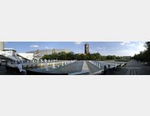 Weitläufiger Platz mit Springbrunnen, umgeben von Bäumen. Im Hintergrund sind ein modernistisches Gebäude und ein historischer Turm sichtbar, daneben Sitzbänke mit Menschen entlang der Promenade.