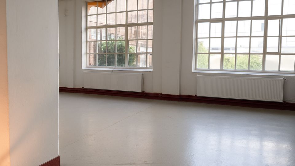 Leerer, heller Innenraum mit weiß gestrichenen Wänden und glattem Boden, geprägt von großen Sprossenfenstern. Unter den Fenstern verlaufen Heizkörper und rote Sockelleisten, draußen sind Gebäude und etwas Grün zu sehen.