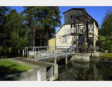 Altes Gebäude einer Wassermühle mit teils verputzten, teils freiliegenden Ziegelwänden und einem Dachstuhl aus Holz; davor eine kleine Brücke und Metallkonstruktionen über einem Wasserkanal, umgeben von Bäumen und Wiesen.