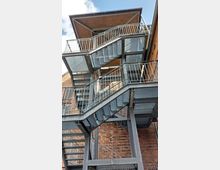 Außentreppe aus Metall mit mehreren Ebenen und Geländern, angebaut an einer Fassade aus rotem Backstein. Im Hintergrund ist ein kleiner überdachter Bereich zu sehen, sowie ein Ausschnitt des Himmels.