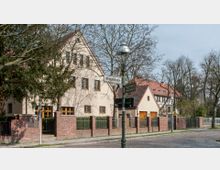 Straßenansicht einer Wohngegend mit hellen Einfamilienhäusern im traditionellen Stil, Ziegeldächern und Sprossenfenstern. Im Vordergrund steht eine Laterne mit Straßenschildern für die „Miquelstraße“ und „Bernadottestraße“, umgeben von Bäumen und einem gepflasterten Gehweg.