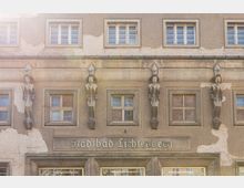 Fassade des Stadtbads Lichtenberg in Berlin mit klassischen Reliefskulpturen von Figuren zwischen den Fenstern. Die Putzstruktur ist stellenweise abgenutzt, und über dem Eingang ist der Schriftzug „Stadtbad Lichtenberg“ zu sehen.