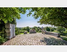 Eine gepflasterte, runde Aussichtsplattform im Grünen mit modernen Steinbänken und einer Skulptur; im Hintergrund dichter Wald und ein weiter Blick über eine Parklandschaft, unter Bäumen.