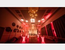 Eleganter Veranstaltungsraum mit roten Lichtakzenten, hohen Decken, einem modernen Kronleuchter und einer erhöhten Bühne im Hintergrund. An den Seiten hängen Vorhänge, und runde Stehtische mit beleuchteten Sockeln sind im Raum verteilt.