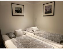 Schlafzimmer mit zwei Einzelbetten, die mit grauen Bettdecken mit Paisleymuster bezogen sind. An der weißen Wand hängen zwei schwarz-weiße Fotografien in Rahmen.