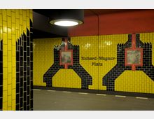 Ein U-Bahnhof mit gelben und schwarzen Kacheln, die geometrische Muster bilden. In der Mitte der Wand steht der Schriftzug „Richard-Wagner-Platz“, flankiert von gerahmten Schwarz-Weiß-Bildern.
