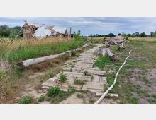 Eine ländliche Szene mit einem provisorischen Holzweg, der durch eine grasbewachsene Fläche führt, daneben umgestürzte Baumstämme und verstreutes Bauholz. Im Hintergrund ist eine teilweise eingestürzte Holzkonstruktion mit einer zerrissenen Plane sichtbar.