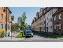 Straßenszene in einem Wohngebiet mit mehrstöckigen Gebäuden auf beiden Seiten, teils aus Ziegel- und Putzfassaden, und roten Satteldächern. Parkende Autos am Straßenrand, Gehweg mit Grünstreifen sowie ein Straßenschild und ein Verkehrsschild im Vordergrund.