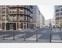 Straßenszene mit modernen Wohn- und Geschäftsgebäuden aus Glas, Stahl und Klinkerfassaden. Im Vordergrund Gehwege mit Pollern sowie eine breite, leere Straße, flankiert von jungen Bäumen und Parkplätzen.