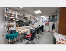 Ein Friseursalon oder Maskenbildnerraum mit mehreren schwarzen Stühlen vor einer langen weißen Arbeitsplatte mit Spiegeln. Regale mit Kosmetikprodukten und Perücken befinden sich an der Wand, während Poster und eine Wanduhr den Raum dekorieren.