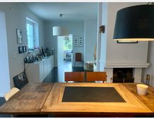 Esszimmer in einem modernen Wohnhaus mit einem Holztisch und Stühlen im Vordergrund, einem weißen Kamin an der rechten Wand und schlichten, grauen Wänden. Im Hintergrund führt ein heller Flur zu weiteren Räumen, neben einem langen Schrank, der mit Familienfotos und Dekorationsgegenständen gestaltet ist.