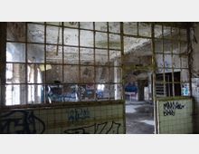 Verlassener, stark beschädigter Raum mit abblätterndem Putz an den Wänden und Decken, durch ein vergittertes Fenster aus der Nähe aufgenommen. Die Wände sind mit Graffiti bedeckt, und auf dem Boden befinden sich Pfützen.