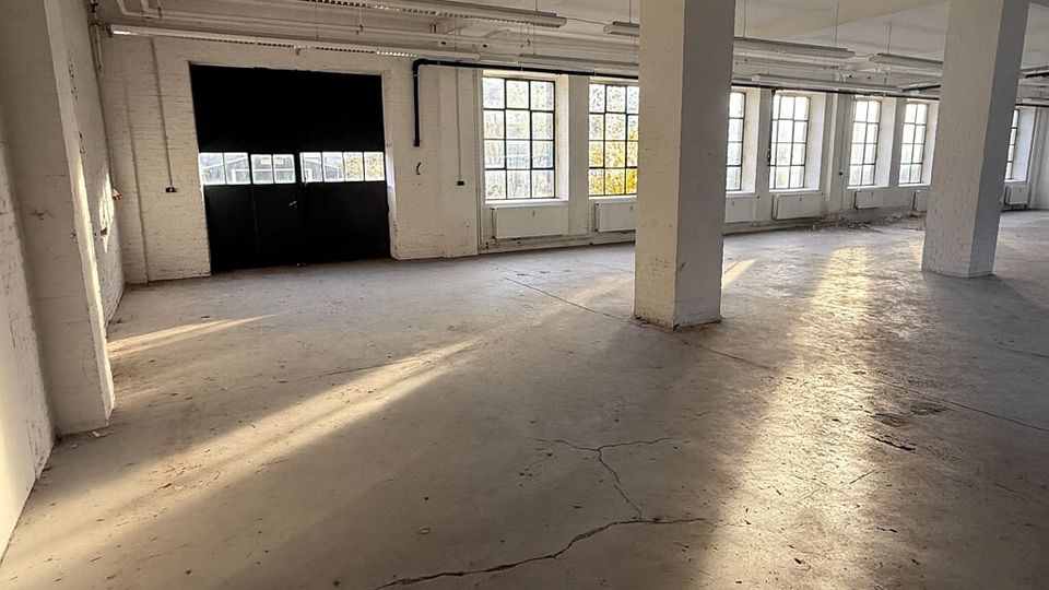 Großer, leerer Industrie- oder Lagerraum mit Betonboden, weißen Stützpfeilern und freiliegenden Deckeninstallationen. An der Seite befinden sich hohe Sprossenfenster und ein großes dunkles Tor, durch die tief stehendes Sonnenlicht lange Schatten wirft.