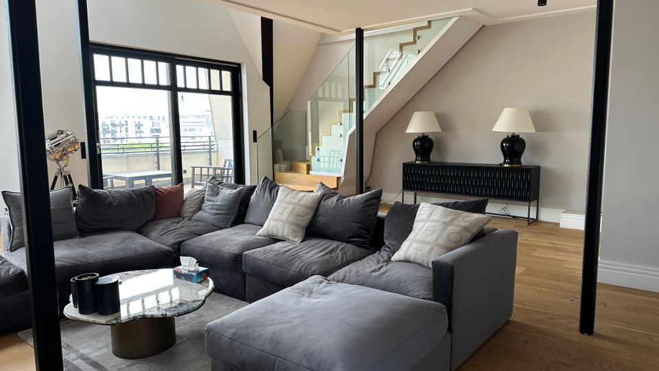 Modernes Wohnzimmer mit großer grauer Eckcouch auf Teppich und rundem Couchtisch vor einer breiten Glasfront mit Zugang zur Terrasse. Im Hintergrund führt eine Treppe mit Holzstufen und Glasgeländer nach oben; daneben stehen ein Sideboard mit zwei Tischlampen und schwarze Stützpfeiler im Raum.