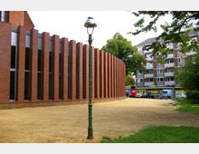 Gebäudekomplex mit einer rotbraunen Ziegelfassade und vertikalen Fensterelementen in abgerundeter Form, umgeben von einer offenen Kiesfläche mit Grünflächen und einem einzelnen Laternenmast im Vordergrund. Im Hintergrund sind Wohngebäude und ein Supermarkt mit geparkten Autos zu sehen.