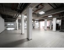 Großer, offener Raum mit Sichtbetondecke, weißen Stützsäulen und hellem Fliesenboden. An der Decke verlaufen sichtbare Rohre, während einzelne Fenster Tageslicht hereinlassen; im Hintergrund sind einige Möbelstücke und Pflanzen zu sehen.