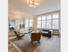 Wohnzimmer in einem hellen Apartment mit großen Fenstern, einem Holzboden im Fischgrätmuster und modernen Möbeln, darunter ein Teppich, ein graues Sofa und zwei Sessel. Im Hintergrund ist ein angrenzendes Esszimmer zu sehen.