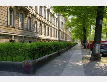 Straßenansicht eines historischen Wohngebäudes mit einer Fassade aus hellen Steinquadern und großen Rundbogenfenstern. Am Gehweg stehen Bäume, die Schatten spenden, während Begrenzungsmauern und eine Hecke den Bereich zwischen Gebäude und Gehweg trennen; parkende Autos befinden sich am rechten Bildrand.