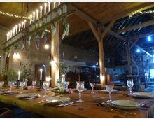 Eine rustikale Scheune mit Holzbalkendecke, geschmücktem Holztisch und hängendem Kerzenleuchter. Der Tisch ist für ein Essen gedeckt, mit Porzellantellern, Gläsern, Besteck und Kerzen, umgeben von dekorativen Blumen.