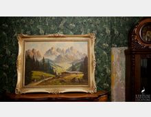 Ein Gemälde in einem reich verzierten, goldenen Rahmen zeigt eine alpenländische Berglandschaft mit Tannen, Hütten und Bergen im Hintergrund. Es steht auf einem dunklen Holztisch vor einer grünen Tapete mit Pflanzenmotiv und neben einem vintage Holzschrank mit Uhr.