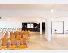 Großer, heller Veranstaltungsraum mit einer Reihe von Holzstühlen, die auf eine schwarze Theke im Hintergrund ausgerichtet sind. Der Raum ist schlicht gestaltet, mit weißen Wänden, hellen Lampen und minimalistischen Dekorationselementen.