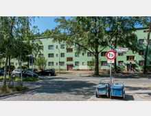 Mehrstöckiges Wohngebäude mit hellgrüner Fassade und roten Fenstern, umgeben von Bäumen und parkenden Autos. Im Vordergrund verläuft eine mit Kopfsteinpflaster belegte Straße, Straßenlaternen und Verkehrsschilder sind zu erkennen.