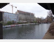 Blick auf einen Kanal mit graffitibedeckter Ufermauer, im Hintergrund mehrstöckige Gebäude, darunter eines mit Baugerüst. Links ragt ein Baukran in den Himmel, Bäume ohne Laub säumen das Ufer.