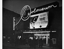 Eine historische Außenaufnahme eines Kinos mit dem Namen „Colosseum“ bei Nacht. Auf der beleuchteten Fassade ist ein großes Filmplakat für „Nackt unter Wölfen“ zu sehen, ergänzt durch eine schlaufenförmige Leuchtschrift. Vor dem Eingang stehen mehrere Menschen, umgeben von gedämpftem Licht.