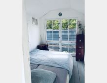 Ein kleines Schlafzimmer mit weißen Wänden und großen Fensterfronten, die Blick auf grüne Bäume und einen Zaun bieten. Im Raum befinden sich ein Bett mit gestreiftem Bettzeug, eine kleine Kommode, eine Blumenvase und eine Holzkiste.