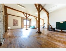 Ein großzügiger, heller Raum mit Holzbalken und Holzboden, teilweise unverputzten Backsteinwänden und großen Fensteröffnungen. Im Raum stehen ein blaues Sofa, ein kleiner Tisch und eine Tafel, ergänzt durch Dekorationen wie ein Gemälde an der Wand.