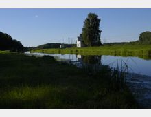 Flusslandschaft mit grasbewachsenen Ufern, einem ruhigen Wasserlauf und einem kleinen weißen Gebäude mit Baum im Hintergrund; umgeben von offenem Feld und bewaldeten Flächen am Horizont.
