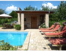 Gartenbereich mit einem Pool, umgeben von einer gepflasterten Terrasse. Links befinden sich ein Esstisch mit Stühlen unter einem Sonnenschirm, rechts stehen mehrere Liegen mit roten Polstern. Im Hintergrund steht ein kleines Gebäude aus Backstein mit einer Holztür und Blumenbeeten.