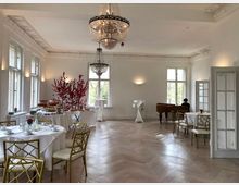 Eleganter Veranstaltungsraum mit großen Fenstern, verzierten Deckenleisten und einem Kronleuchter. Im Raum stehen weiß gedeckte Tische mit goldenen Stühlen, ein Buffet mit roten Dekorationen, und im Hintergrund ein Flügel, an dem eine Person sitzt.