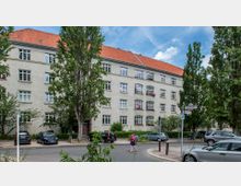 Wohngebäude mit heller Fassade und roten Ziegeldach, umgeben von Bäumen und parkenden Autos. Im Vordergrund ist eine Straße mit einem Verkehrsschild und einer Passantin zu sehen.