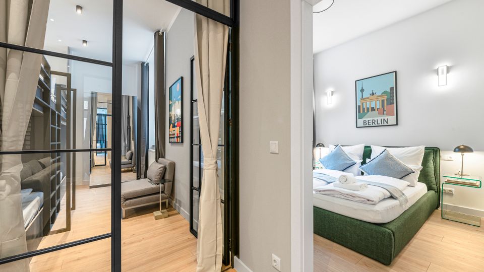 Heller, modern eingerichteter Apartmentflur mit Glas-Metall-Trennwand und bodentiefen Vorhängen, Holzboden und Sitzbereich. Rechts öffnet sich ein Schlafzimmer mit grünem Polsterbett, Nachttischen und einer „Berlin“-Wandgrafik.