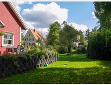 Garten mit Rasenfläche, Bäumen und Sträuchern, umgeben von Einfamilienhäusern, darunter ein rotes Holzhaus links im Bild mit Terrasse und Gartenmöbeln. Der Himmel ist teilweise bewölkt, und herbstliche Blätter liegen verstreut auf dem Rasen.