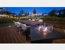 Holzterrasse in einem Garten bei Dämmerung mit großem Esstisch und mehreren weißen Stühlen sowie einem modernen Gas-Feuertisch im Vordergrund. Im Hintergrund sind bepflanzte Beete, Lichterketten, eine Hängematte unter einer Pergola und Wohnhäuser zu sehen.