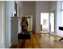 Ein heller Flur mit Holzdielenboden, rechts ein großer Wandspiegel und links ein Heizkörper mit Dekoration. Im Hintergrund führen Türen in ein weiteres Zimmer mit Holzmöbeln und einem Teppich.