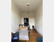 Schmaler Raum mit hellem Holzboden, einem Einzelbett links und einer dunklen Regalwand dahinter. Im Hintergrund ist ein offener Kleiderschrank zu sehen, an der Decke hängt eine filigrane schwarze Pendelleuchte.