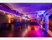 Industrieartige Veranstaltungslocation mit weiß gewölbter Decke, bunten Lichteffekten und Discokugeln. Der Raum ist mit Stehtischen und Hockern ausgestattet, während die Wände durch orangefarbene und blaue Beleuchtung akzentuiert werden.