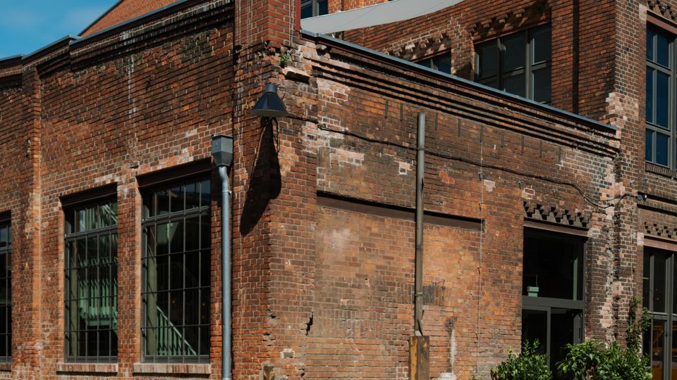 Red-brick industrial-style building with tall multi-pane windows and weathered masonry, set along a paved courtyard. A glass entryway with potted plants sits at the corner beneath a sloped roofline and upper-level windows.