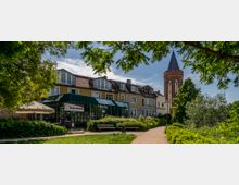 Eine malerische Szene mit einem gelben Gebäude, das moderne und traditionelle Architektur kombiniert, einschließlich eines Wintergartens und Terrassenbereichs mit Sonnenschirmen, flankiert von einem Gehweg mit Bänken und üppiger Begrünung. Im Hintergrund ist ein roter Backsteinturm mit spitzem Dach zu sehen, vermutlich Teil einer Kirche.