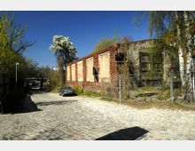 Pflasterstraße mit einem geparkten Auto neben einem alten, teilweise verfallenen Ziegelgebäude. Im Hintergrund sind eine Brücke, blühende Bäume und ein klarer blauer Himmel zu sehen.