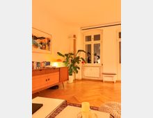 Helles Wohnzimmer mit Holzparkett und großem Doppelfenster über einem Heizkörper. Links stehen ein Mid-Century-Sideboard mit Büchern und Lampe sowie eine große Zimmerpflanze; im Vordergrund sind ein niedriger Tisch mit Kerze und ein gemusterter Teppich zu sehen.