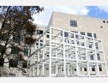 Moderne Gebäudeansicht mit einer hellen Fassade aus rechteckigen Steinplatten und großen, symmetrischen Fenstern. Im Vordergrund steht eine weiße, kubische Raumskulptur aus Metallstreben vor einem leicht bewölkten Himmel; links ist ein Baum mit braunem Laub sichtbar.